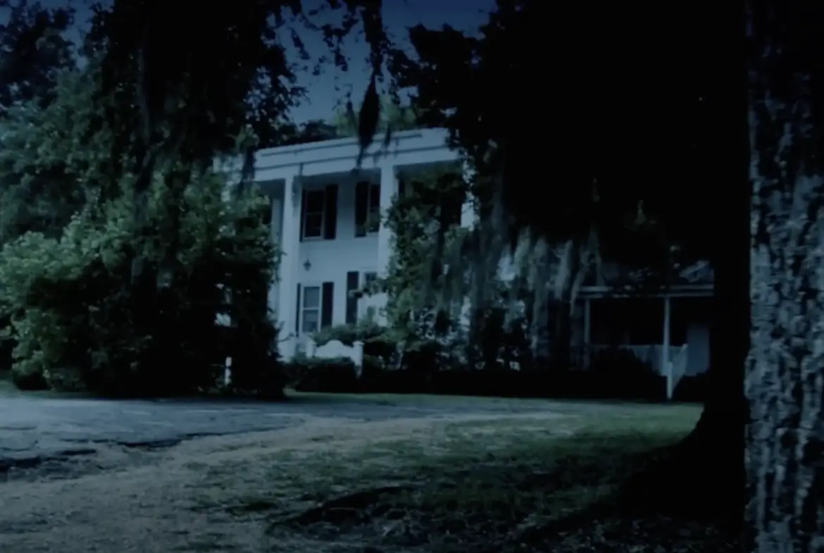 Nighttime view of a historic white two-story Southern mansion, partially obscured by trees and draped with Spanish moss, evoking a mysterious atmosphere.